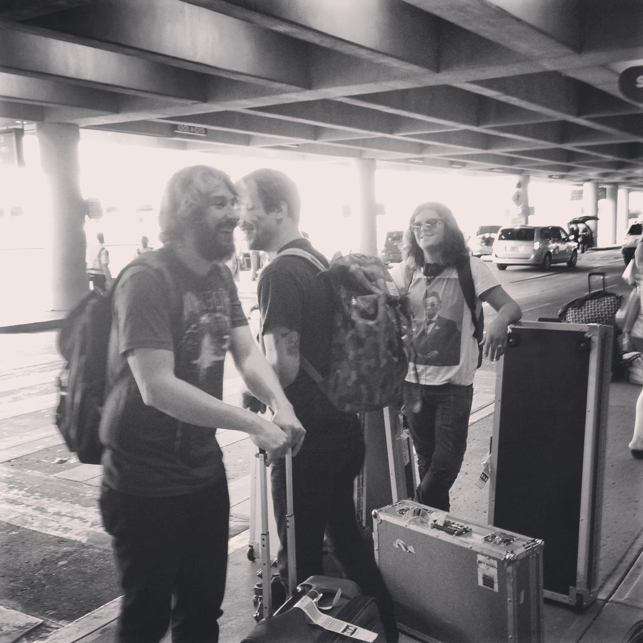 Three unidentified Wild Feathers members standing with instrument and road cases at an airport curb.