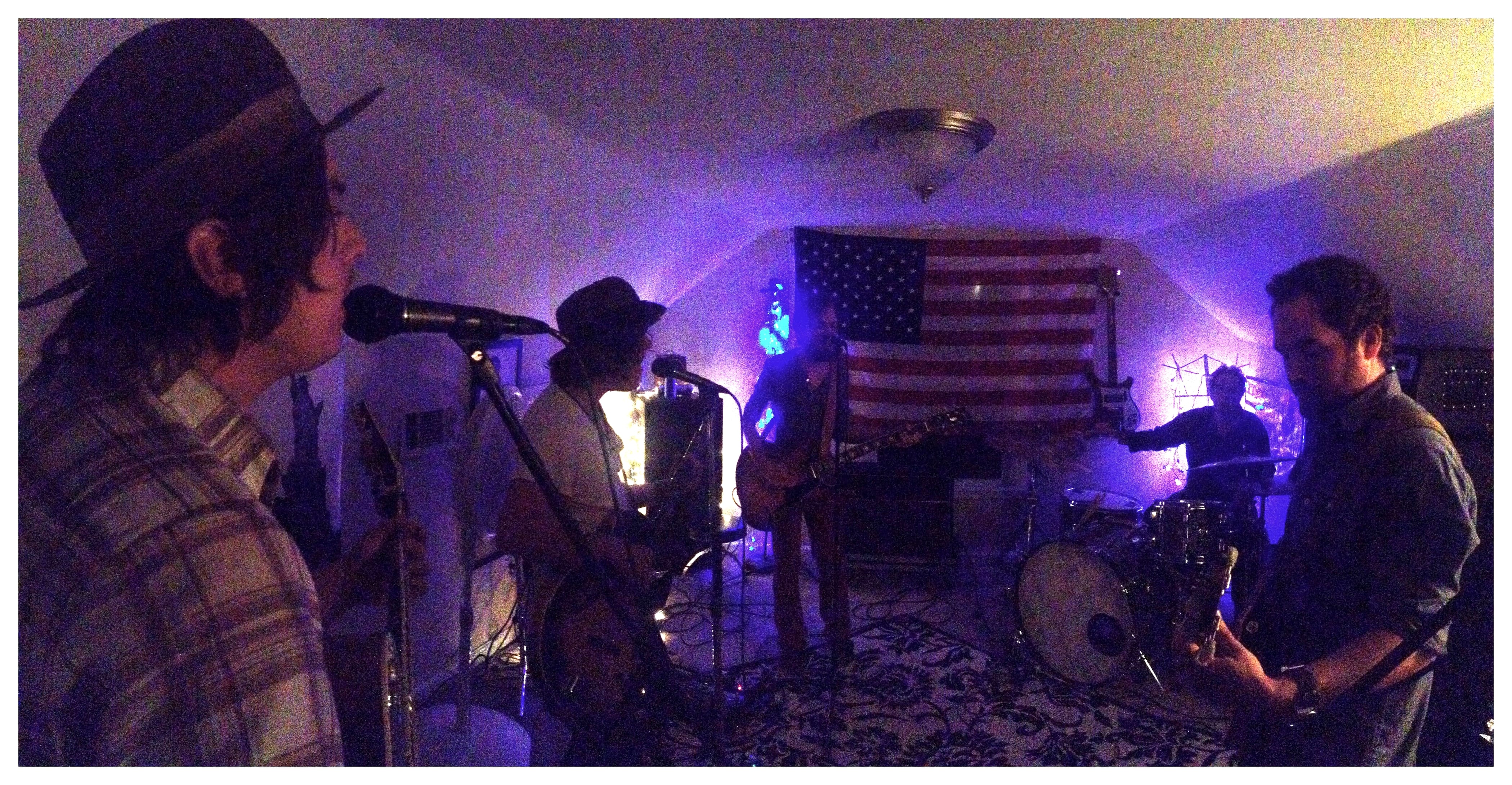 Five musicians rehearse with guitars, drums, and microphones in a low-lit room with an American flag behind them.