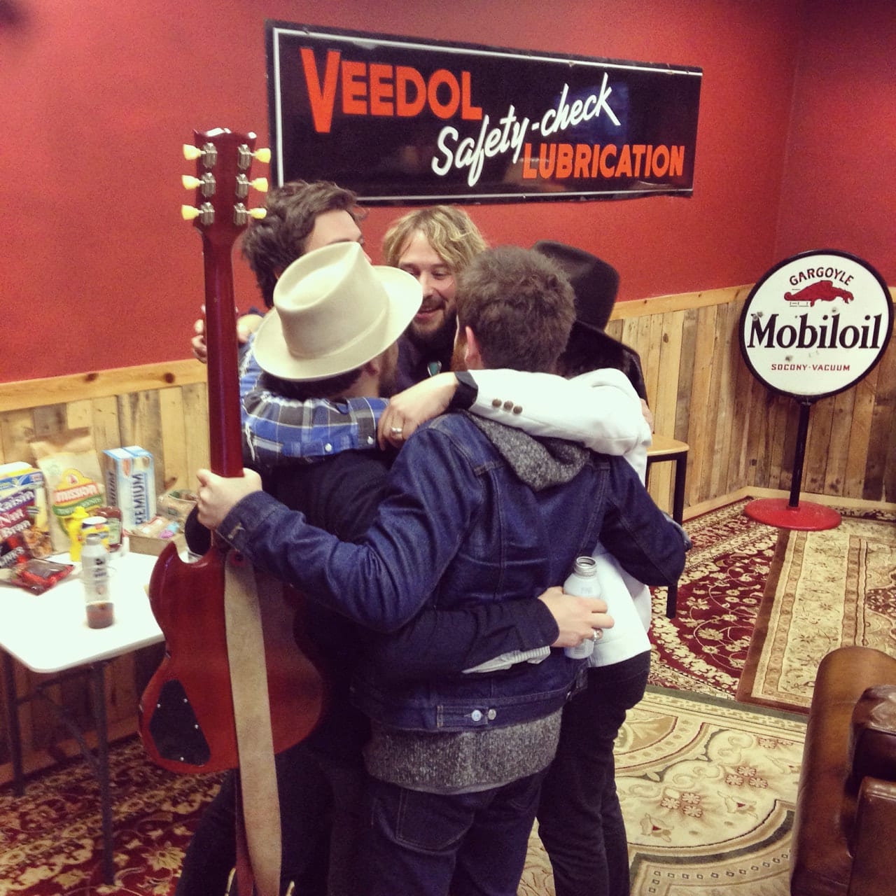 Members of The Wild Feathers embracing in a vintage gas-station-themed lounge or dressing room.