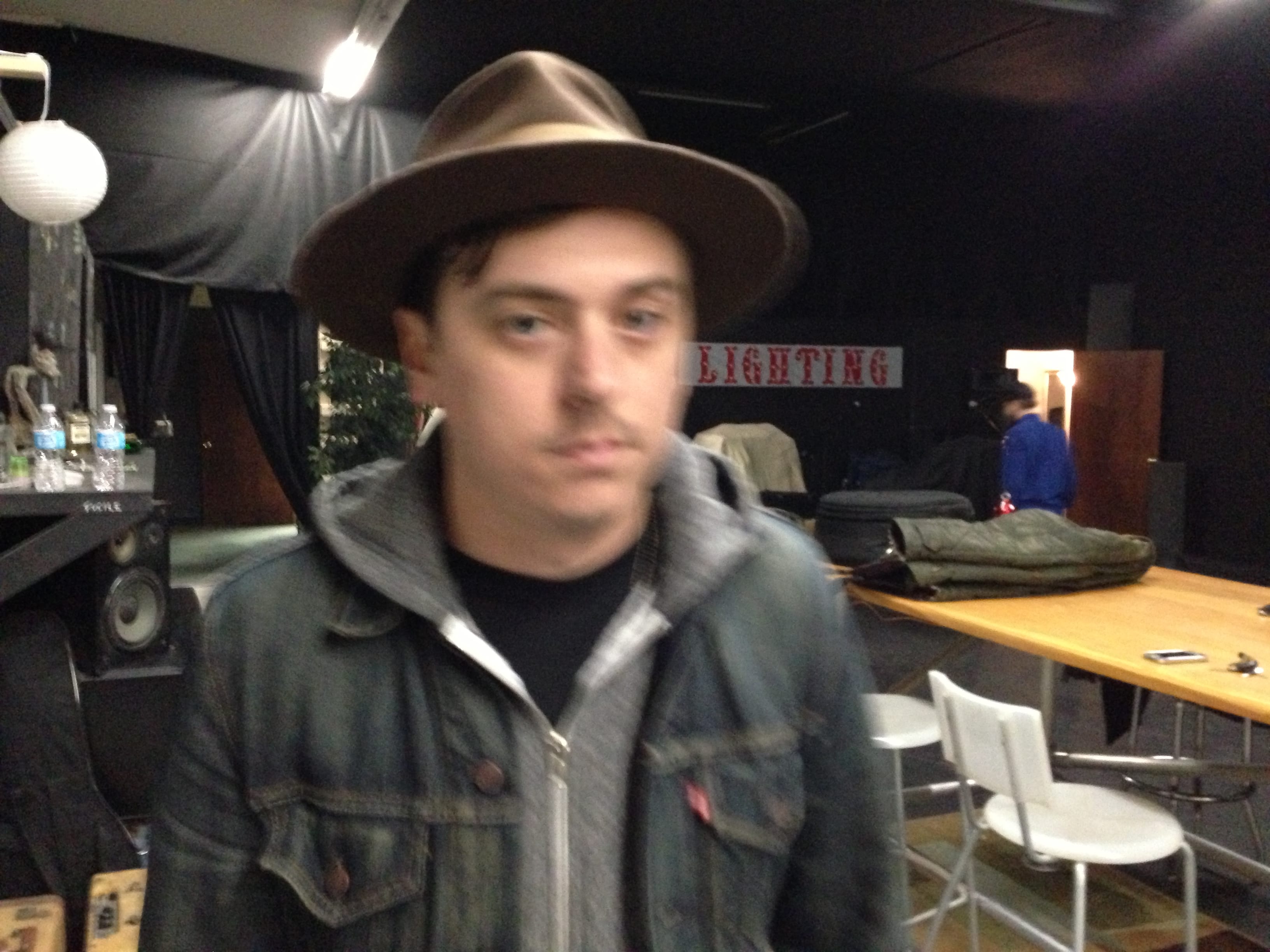 A slightly blurred portrait of an unidentified Wild Feathers member in a hat and denim jacket inside a backstage or production room marked LIGHTING.