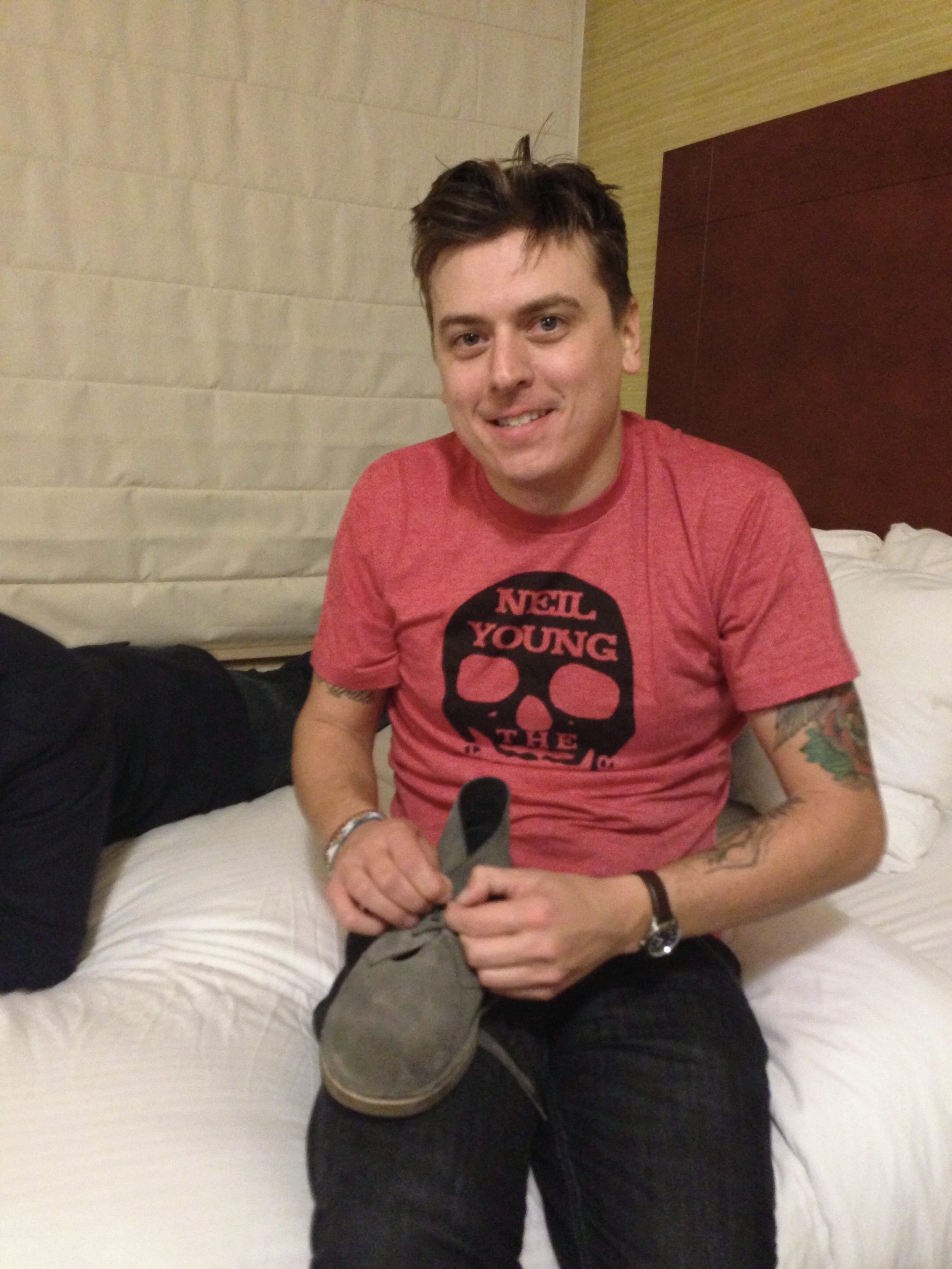 An unidentified member of The Wild Feathers sitting on a hotel bed in a red Neil Young shirt while holding a gray shoe.