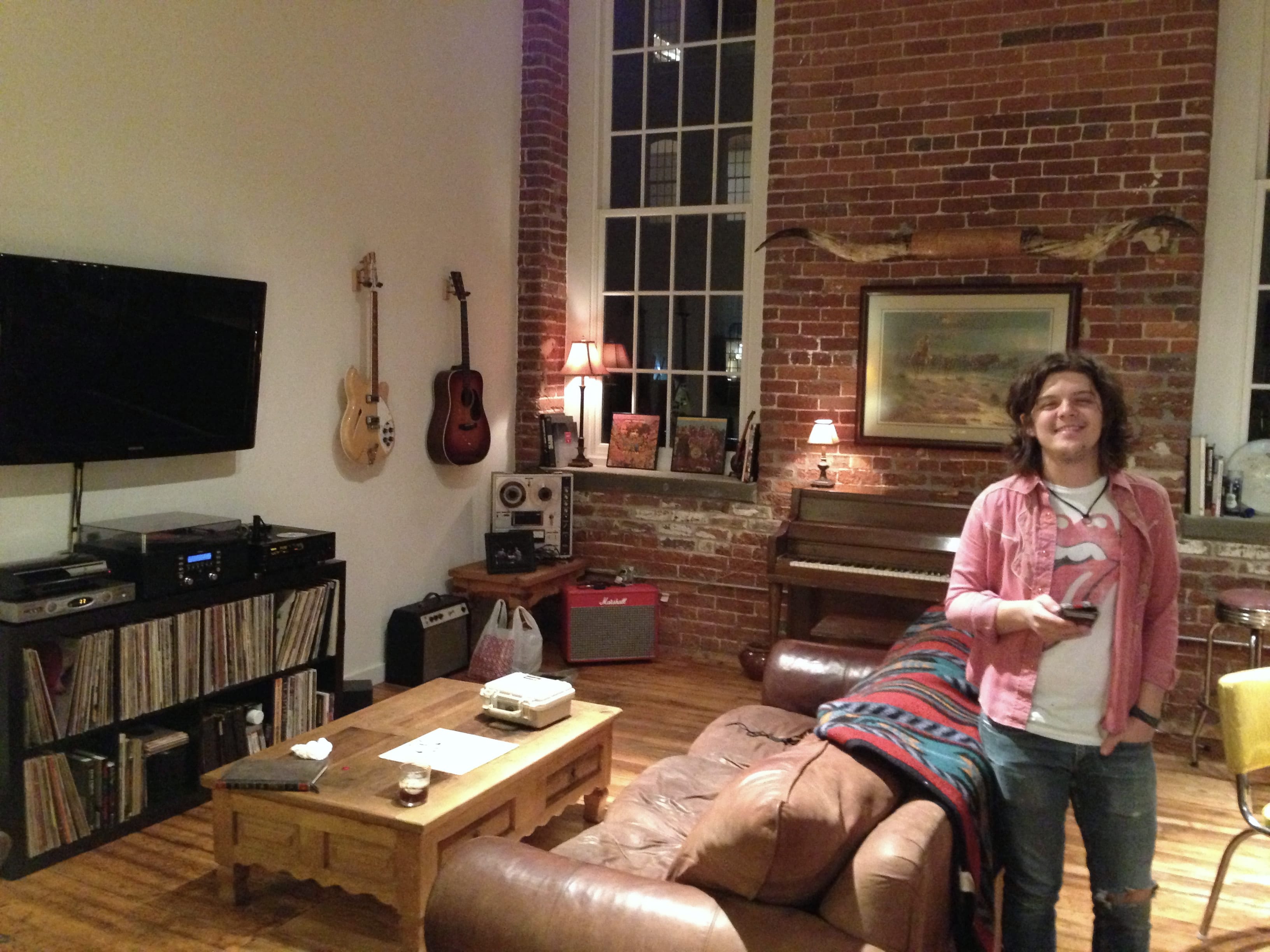 An unidentified member of The Wild Feathers standing in a brick-walled living room with guitars, records, amplifiers, a piano, and a reel-to-reel machine.
