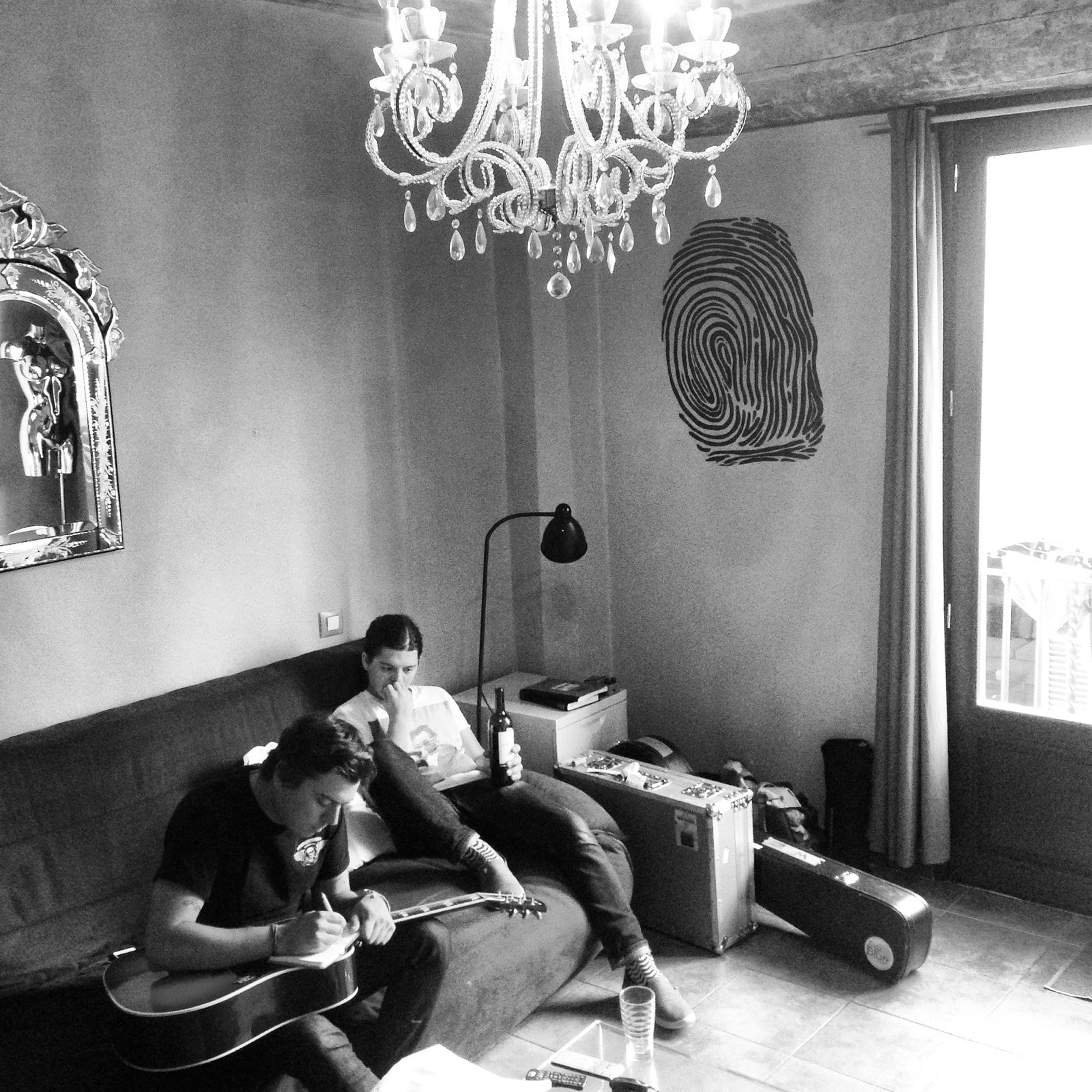A black-and-white photograph shows one person working with an acoustic guitar and notebook on a Barcelona apartment couch while the founding guitarist sits beside them holding a bottle.