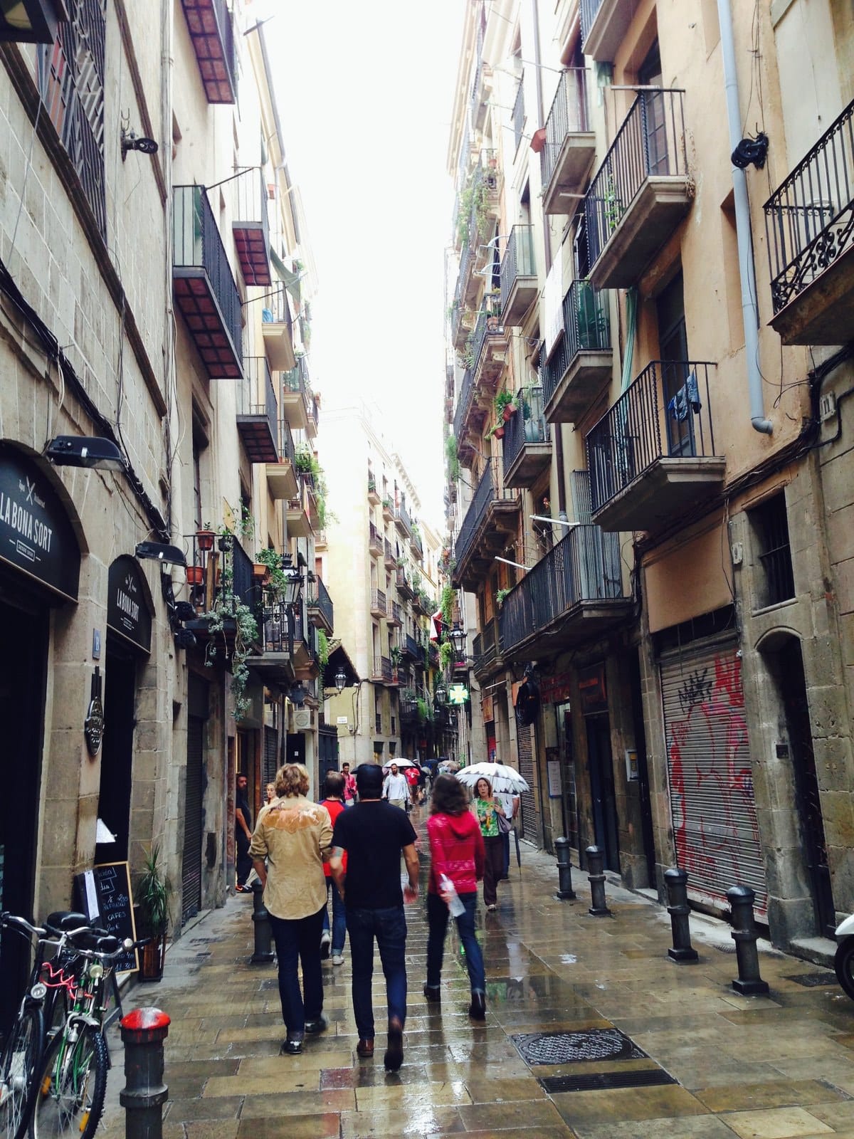 Several people walk down a narrow Barcelona street lined with balconies and parked bicycles.