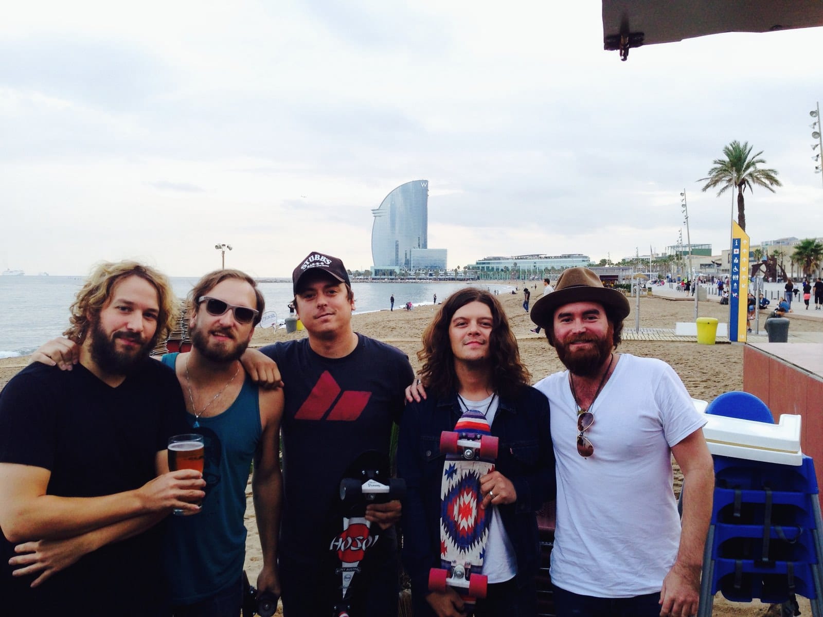 Five members of The Wild Feathers pose at Barceloneta Beach with the W Barcelona hotel visible in the distance.
