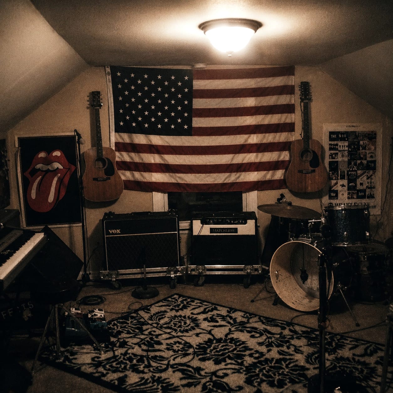 A small rehearsal room with guitars, amplifiers, drums, keyboards, an American flag, and music posters.