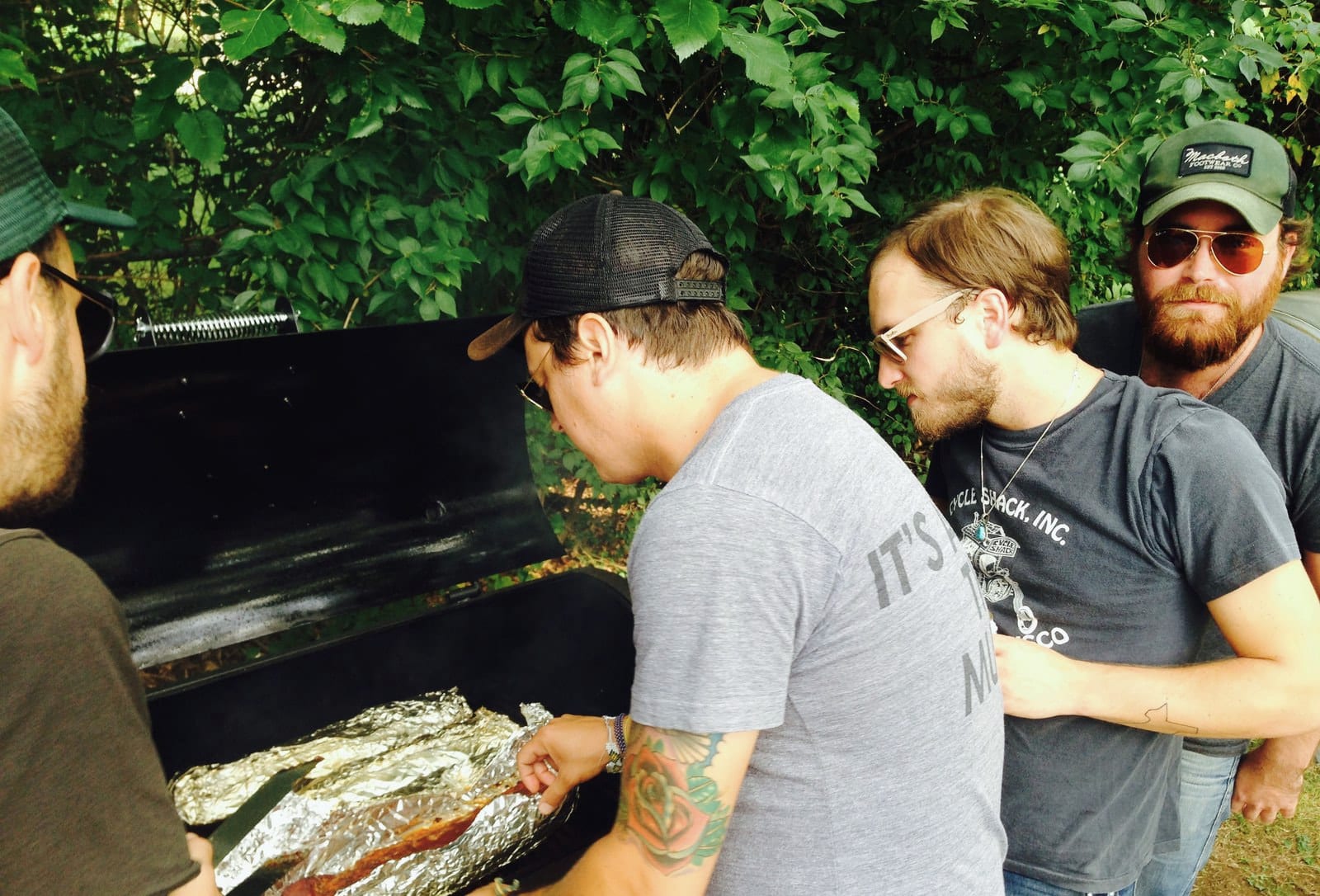 Several people gather around an outdoor grill covered with foil-wrapped fish.