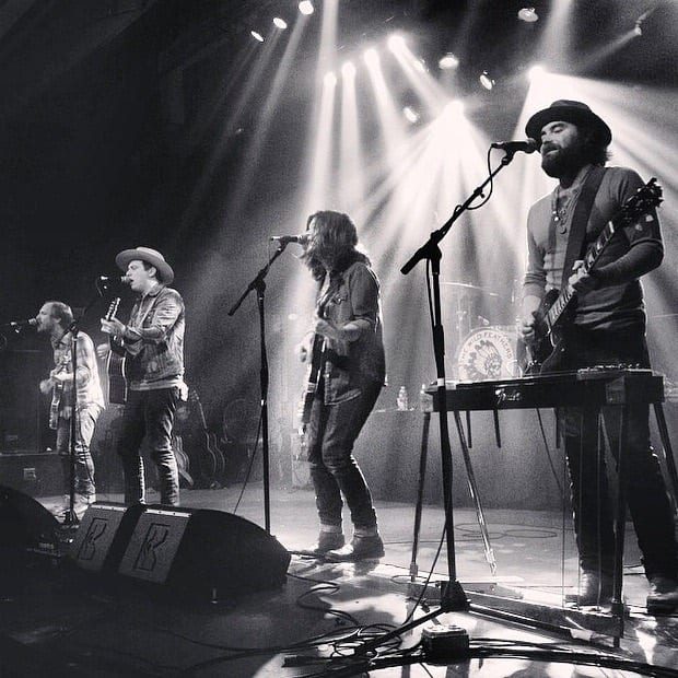 Black-and-white live photograph of four unidentified Wild Feathers members singing and playing across a concert stage.