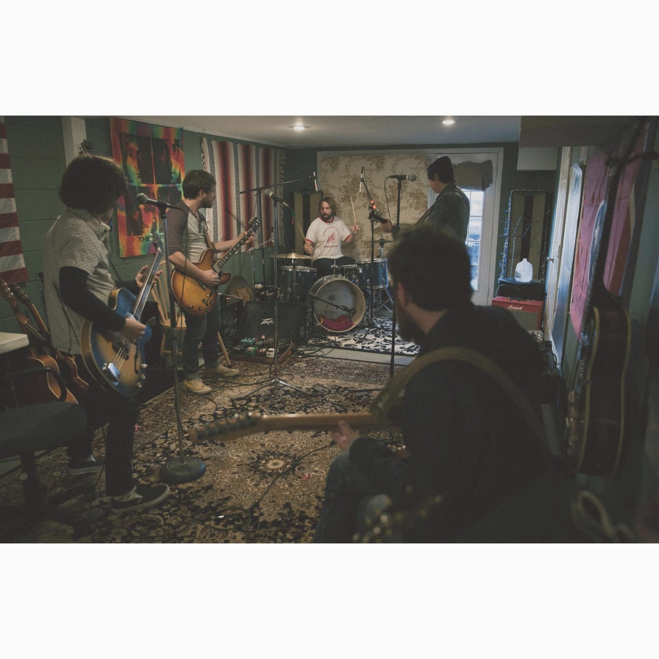 Five unidentified members of The Wild Feathers rehearsing with guitars and drums in a rug-lined room.