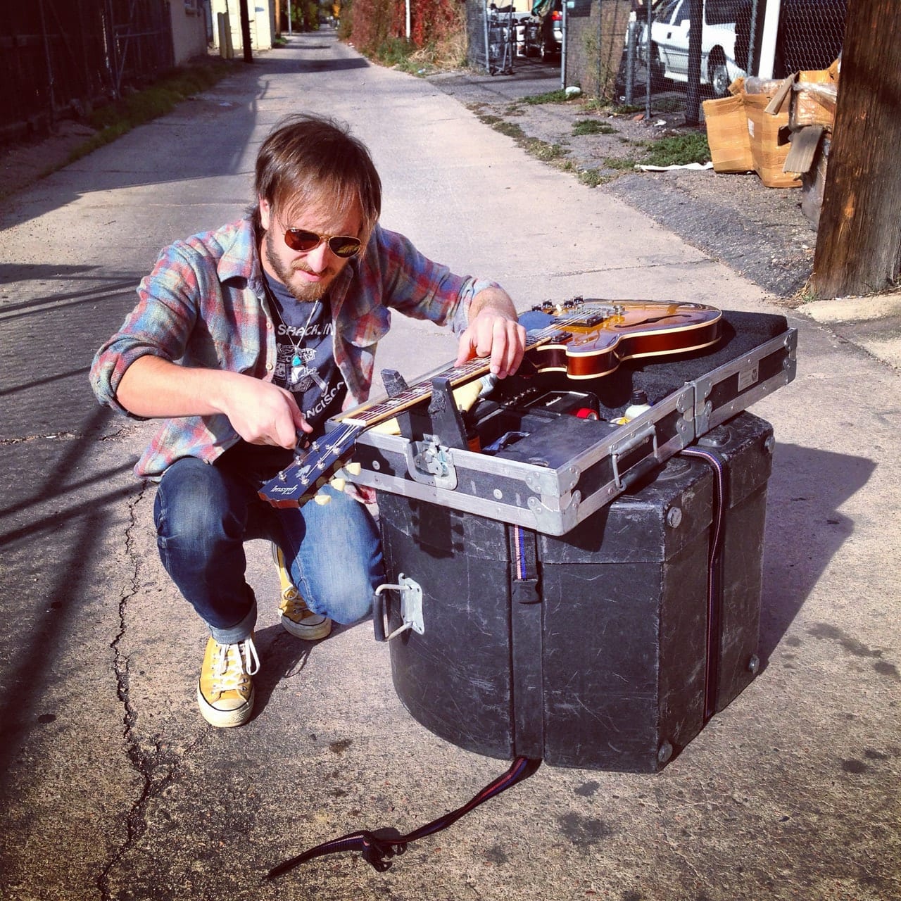 Unidentified person crouched beside road cases while working on a guitar in a sunlit alley.