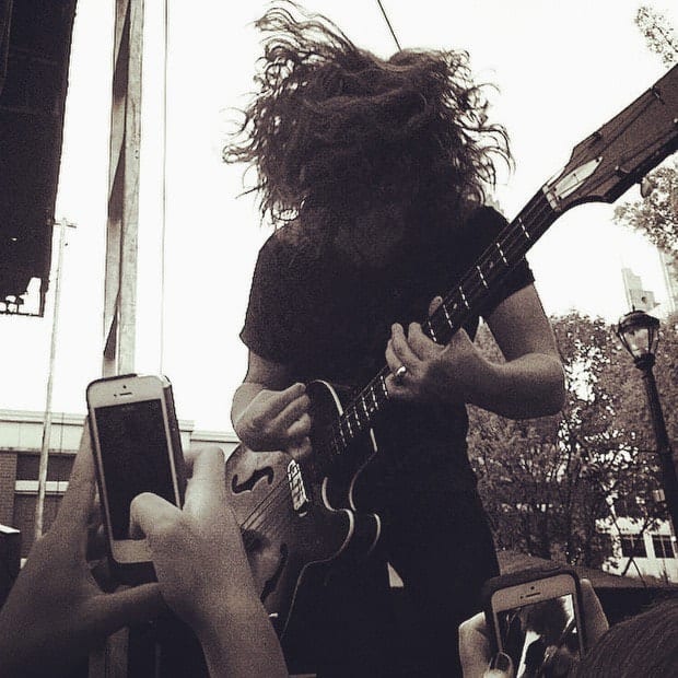 An unidentified Wild Feathers guitarist performing at the edge of an outdoor stage as audience members hold up phones.
