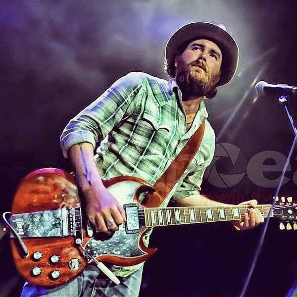 Unidentified Wild Feathers guitarist in a hat and plaid shirt playing an SG-style electric guitar onstage.