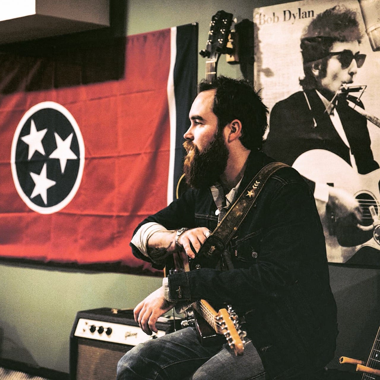 Unidentified bearded band member seated with an electric guitar between a Tennessee flag and a Bob Dylan poster.