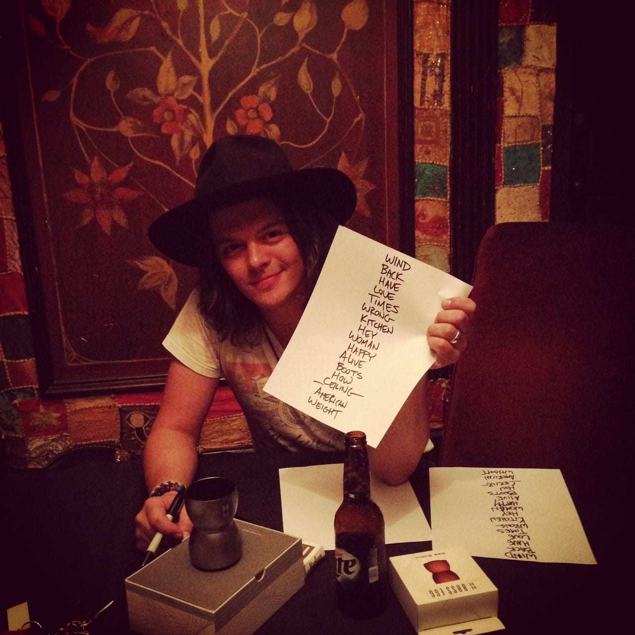 An unidentified Wild Feathers member holding a handwritten set list at a table with a bottle, cup, marker, and another song list.