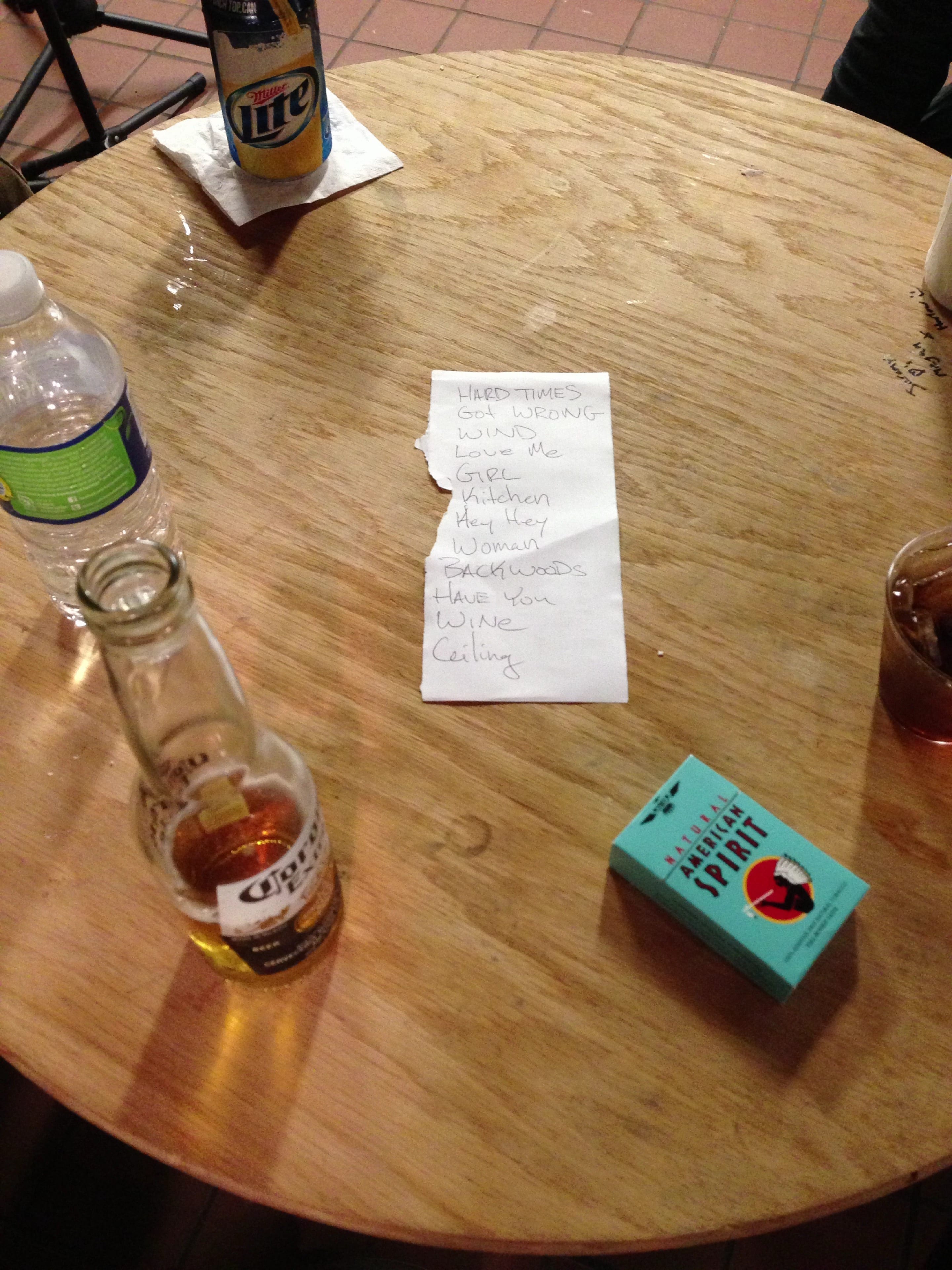 Handwritten set list on a round wooden table among bottles, a water bottle, a drink, and a pack of Natural American Spirit cigarettes.