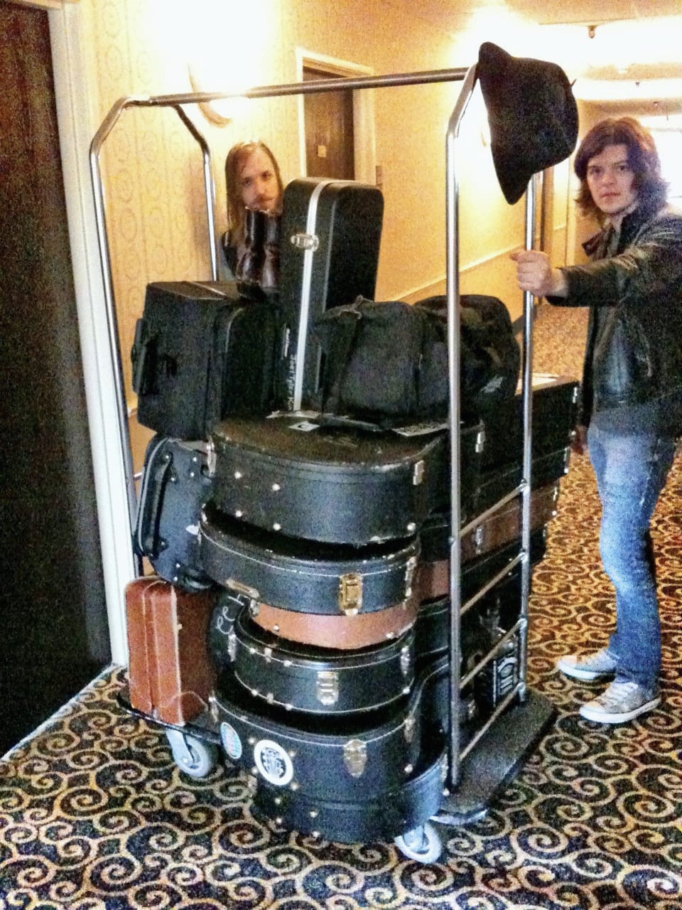 Two people maneuvering a hotel luggage cart stacked with guitars, cases, bags, and a hat.
