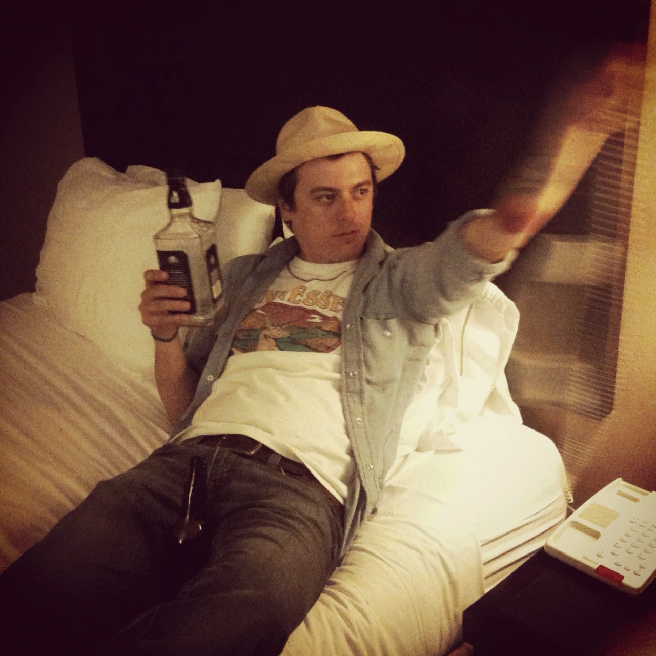 Unidentified band member in a hat reclining on a hotel bed and gesturing while holding a beverage container.