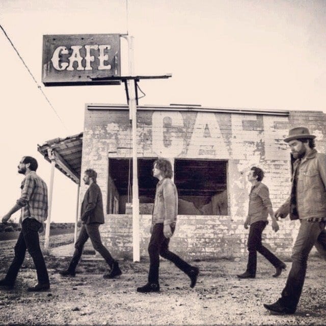 A monochrome Wild Feathers promotional image showing five band members walking past a roadside cafe.