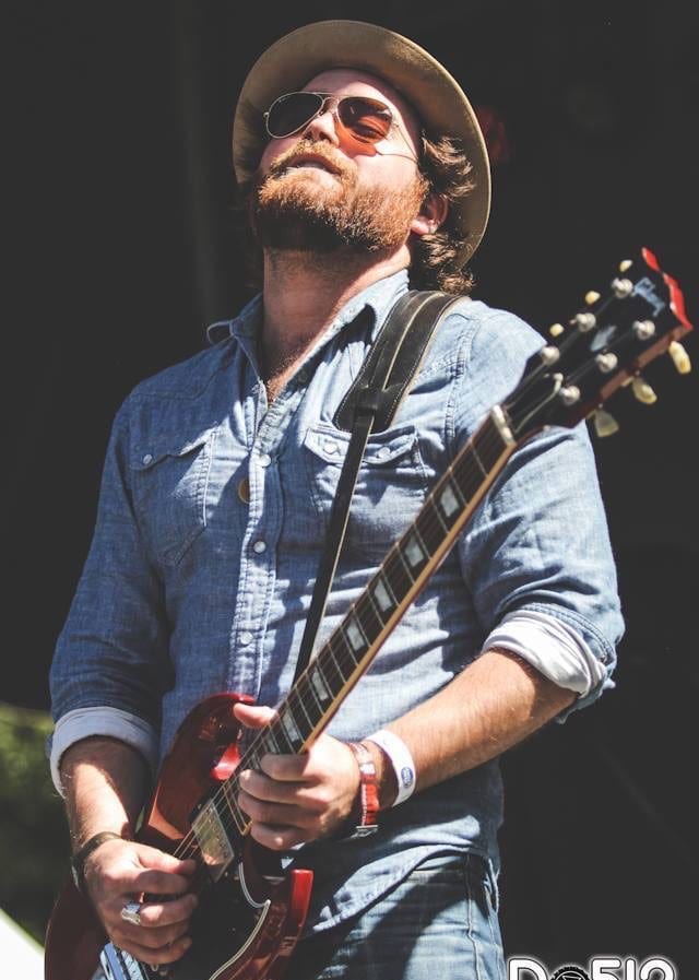 Unidentified Wild Feathers guitarist in a hat and sunglasses playing a red electric guitar onstage.