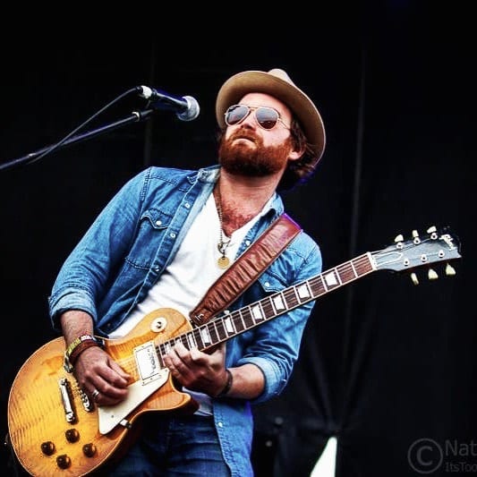 Unidentified Wild Feathers guitarist in sunglasses and a hat playing a gold-finish electric guitar onstage.