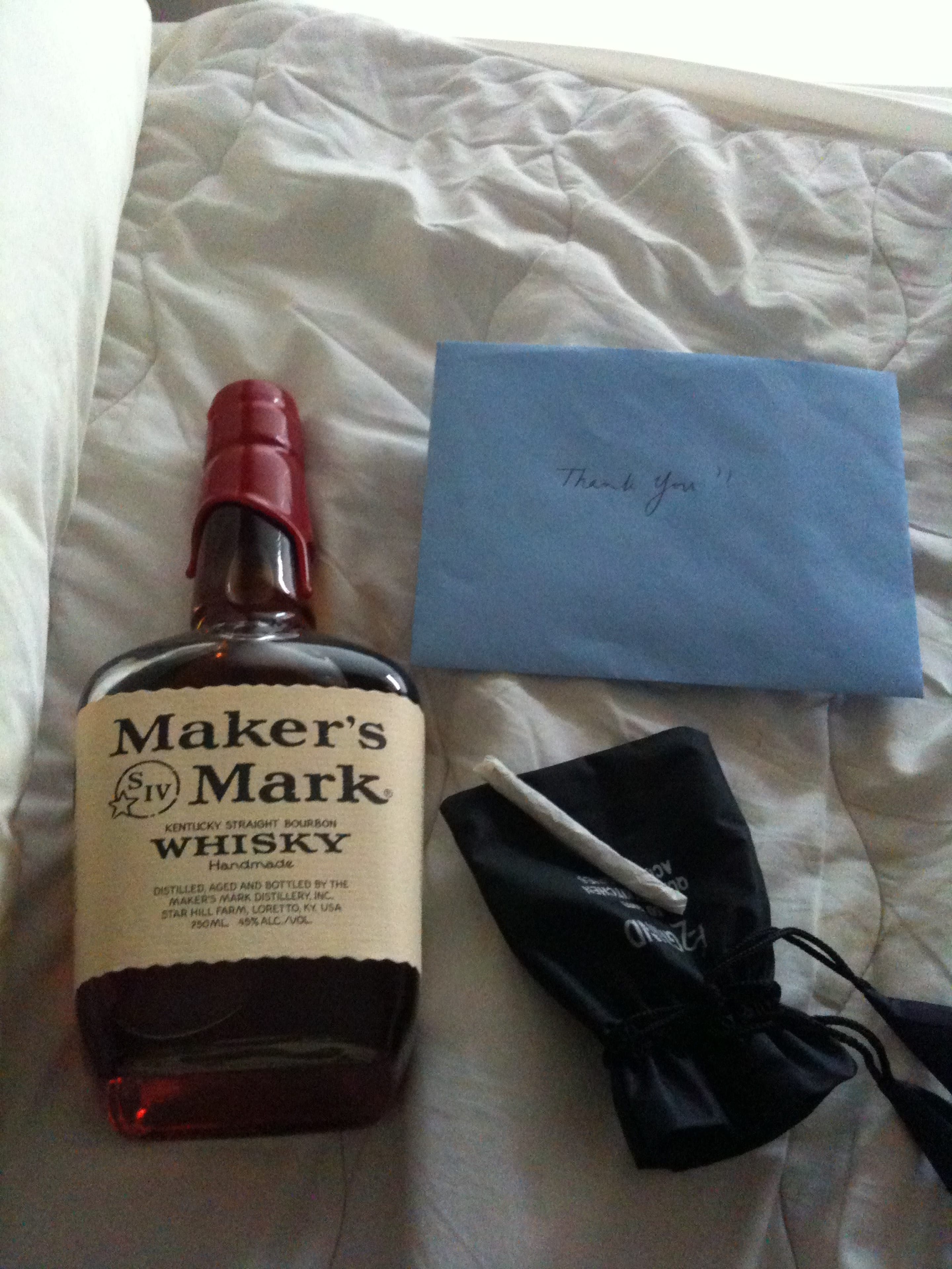 Bottle of Maker's Mark, a blue envelope marked Thank You, and a dark pouch arranged on a hotel bed.