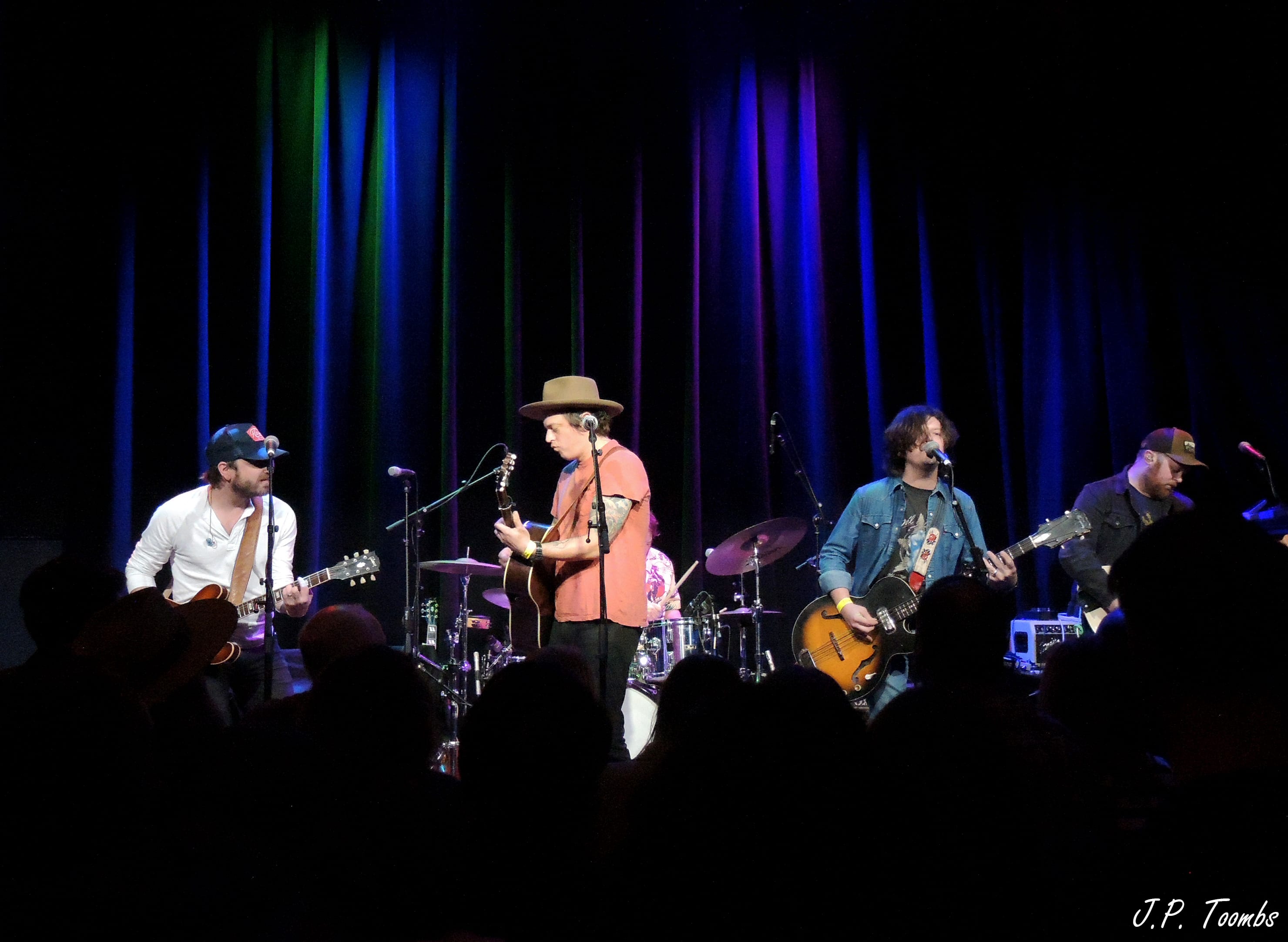 Four members of The Wild Feathers perform across the dark stage at Natalie's Grandview beneath blue and violet light.