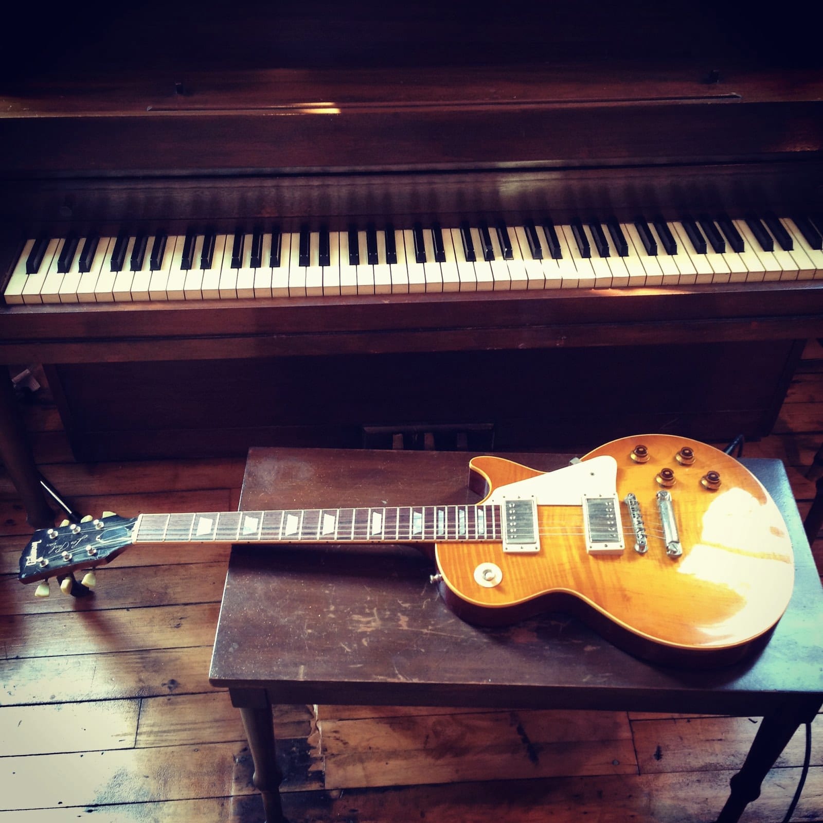 A sunburst electric guitar rests on a wooden floor beneath an upright piano.