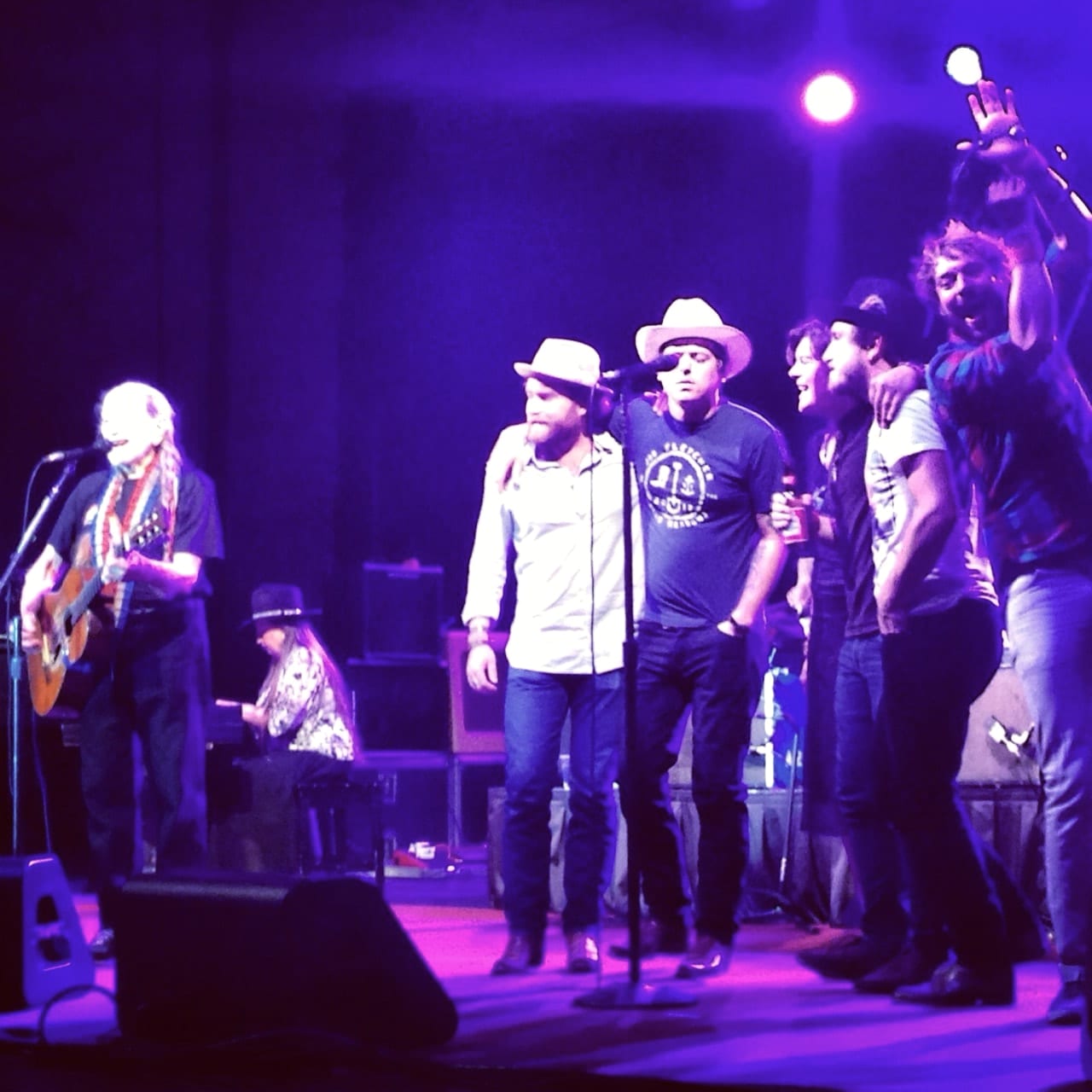Willie Nelson performing at left while members and associates of The Wild Feathers gather around a center microphone under purple stage lights.