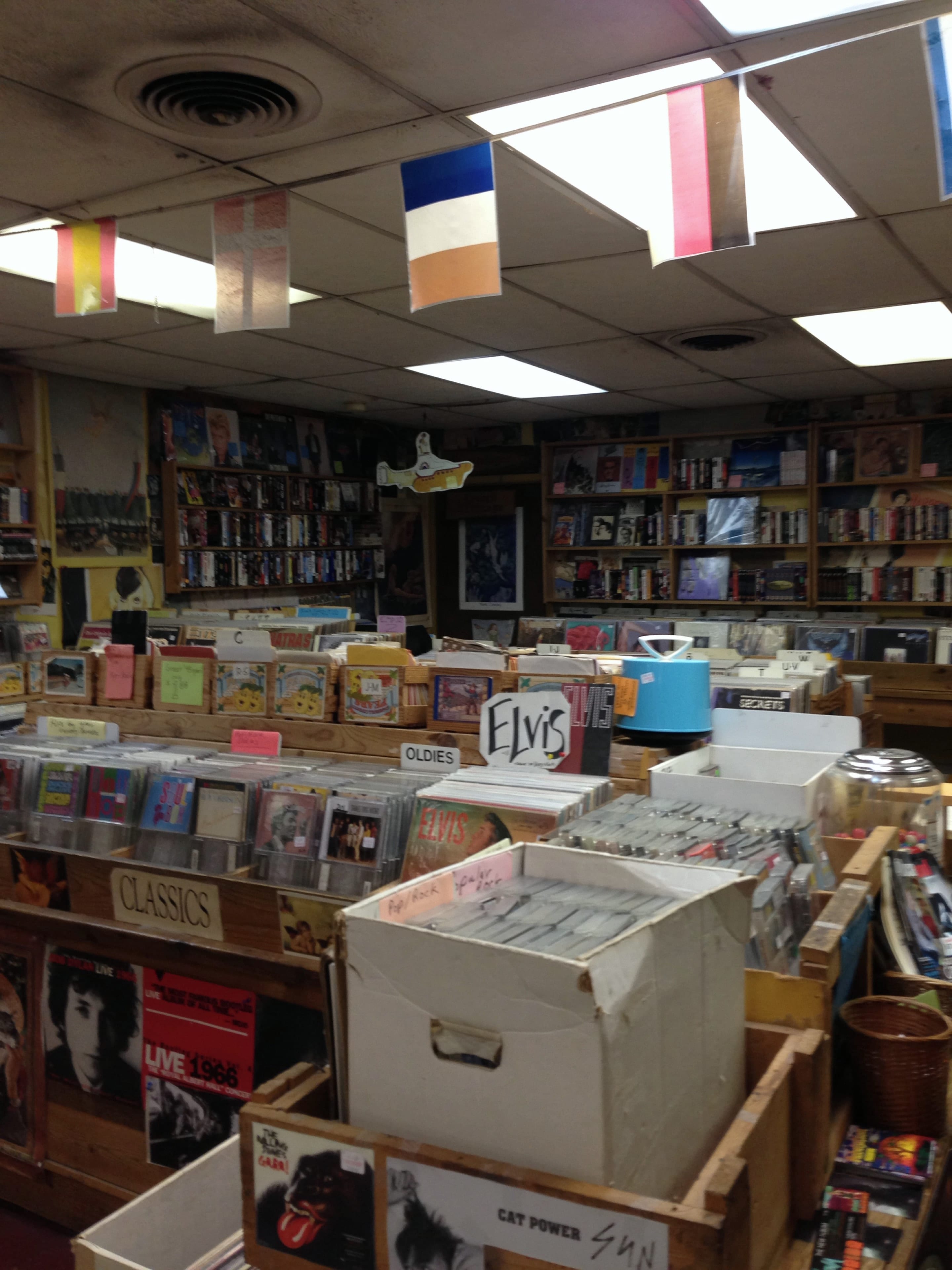 A record-store interior with wooden vinyl bins, handwritten Oldies, Classics, and Elvis labels, bookshelves, and colored signal flags overhead.