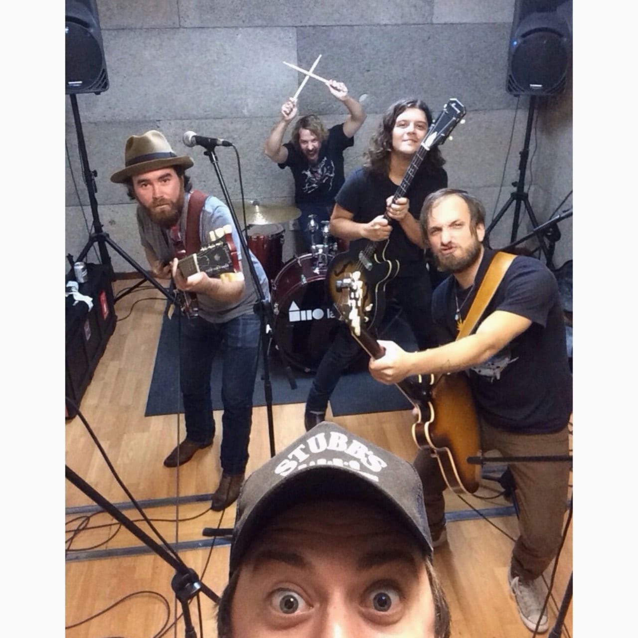 One unidentified person taking a close selfie while four Wild Feathers members pose with guitars and drums in a rehearsal room.