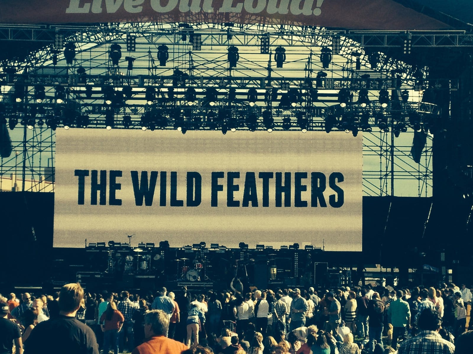 A large stage screen reading The Wild Feathers above a crowd at Reunion Park in Dallas.