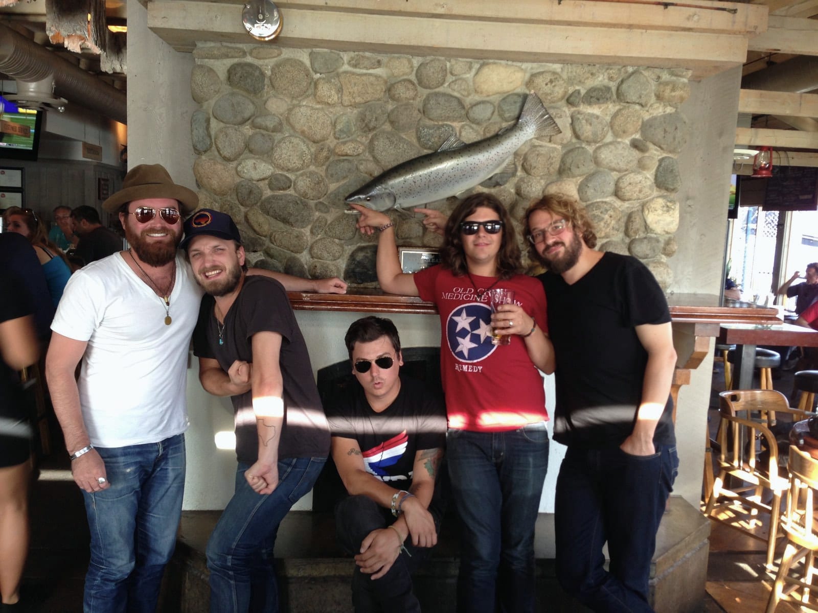 Five band members pose around a stone fireplace and mounted fish inside a restaurant.