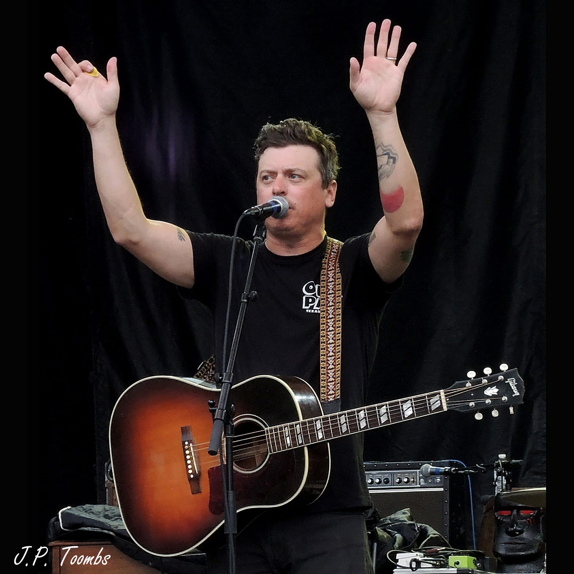 A Wild Feathers member stands behind an acoustic guitar and raises both hands toward the Rock the Ruins audience.