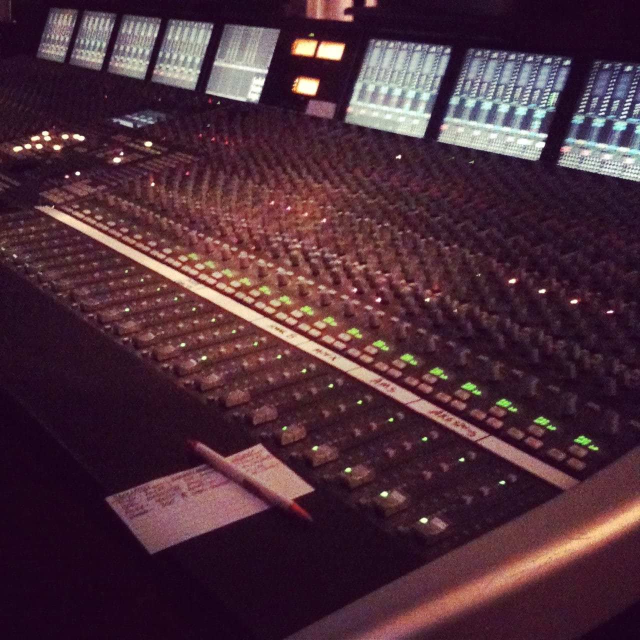 Large recording-studio mixing console with illuminated channel controls, computer displays, a notepad, and pencil.