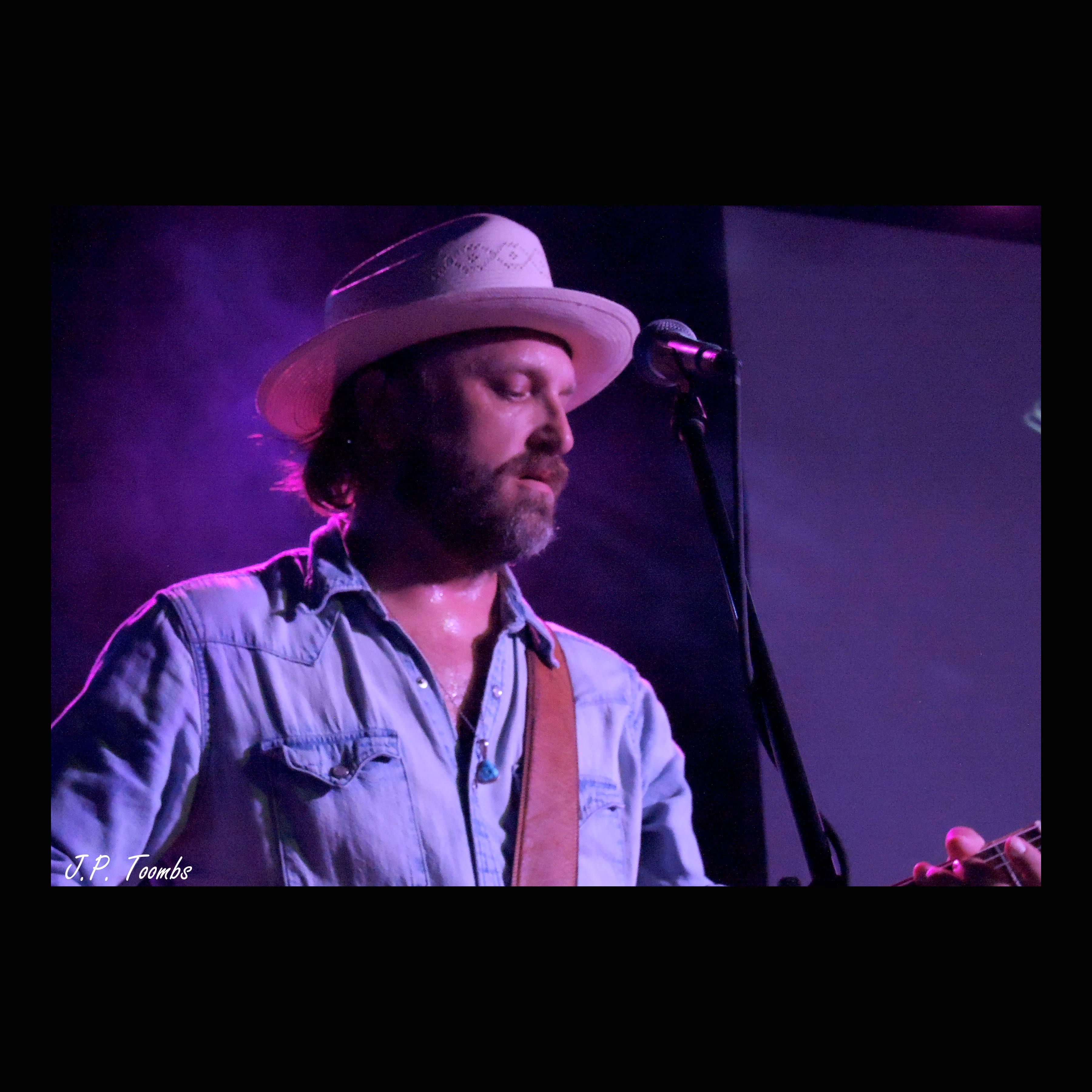 A bearded Wild Feathers guitarist in a light hat sings into a microphone under violet stage light at 3rd & Lindsley.
