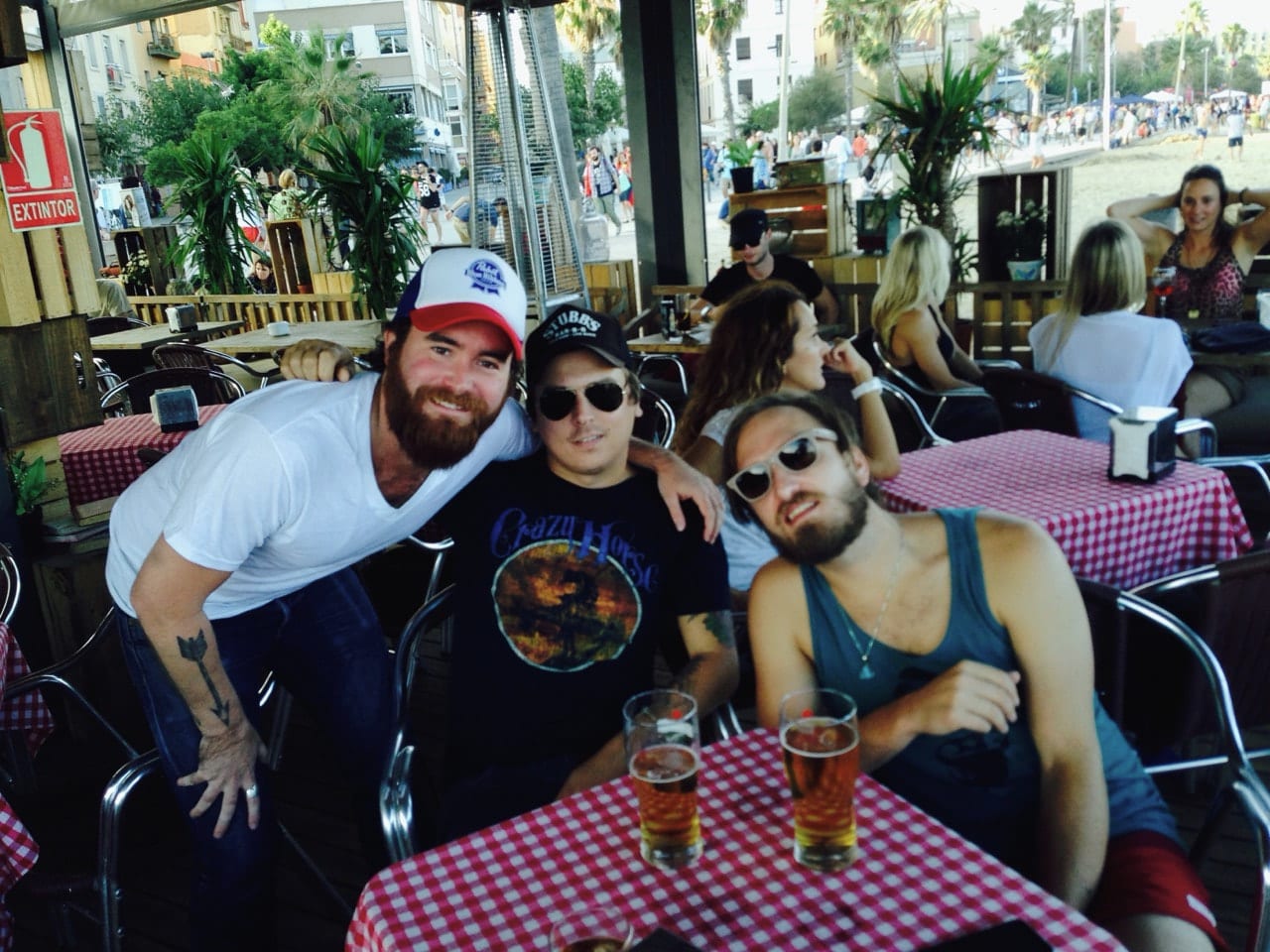 Three unidentified Wild Feathers members seated at an outdoor café with red gingham tablecloths near a palm-lined waterfront.