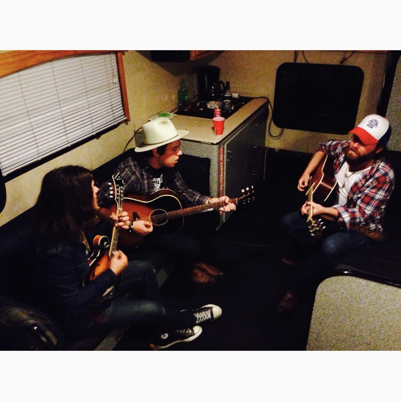 Three unidentified musicians seated inside a bus or trailer and playing two acoustic guitars and a mandolin.