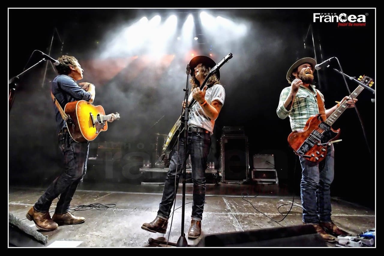 Three unidentified Wild Feathers members singing and playing guitars together on a concert stage.