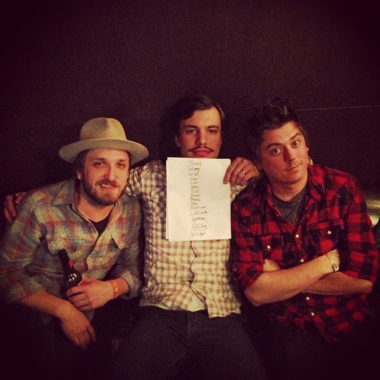 Three unidentified people seated together in a dim room, with the person in the center holding a handwritten set list.