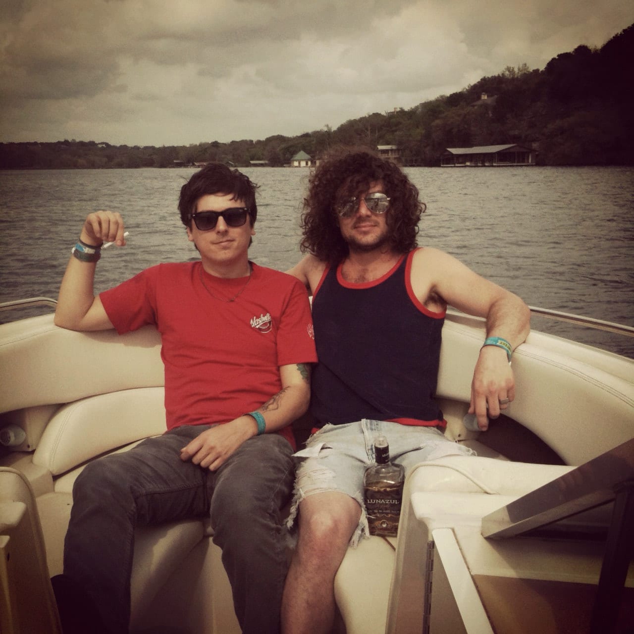 Two unidentified band members wearing sunglasses seated together on a boat with a wooded shoreline behind them.