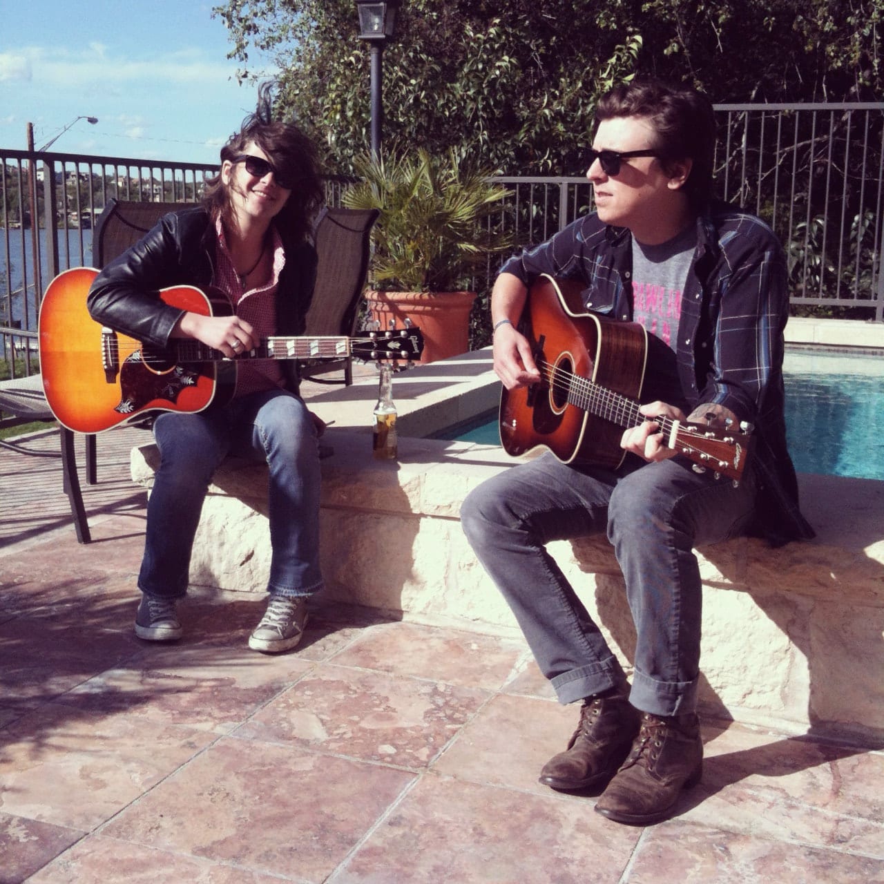 Two unidentified Wild Feathers members wearing sunglasses and playing acoustic guitars beside a swimming pool near Lake Austin.