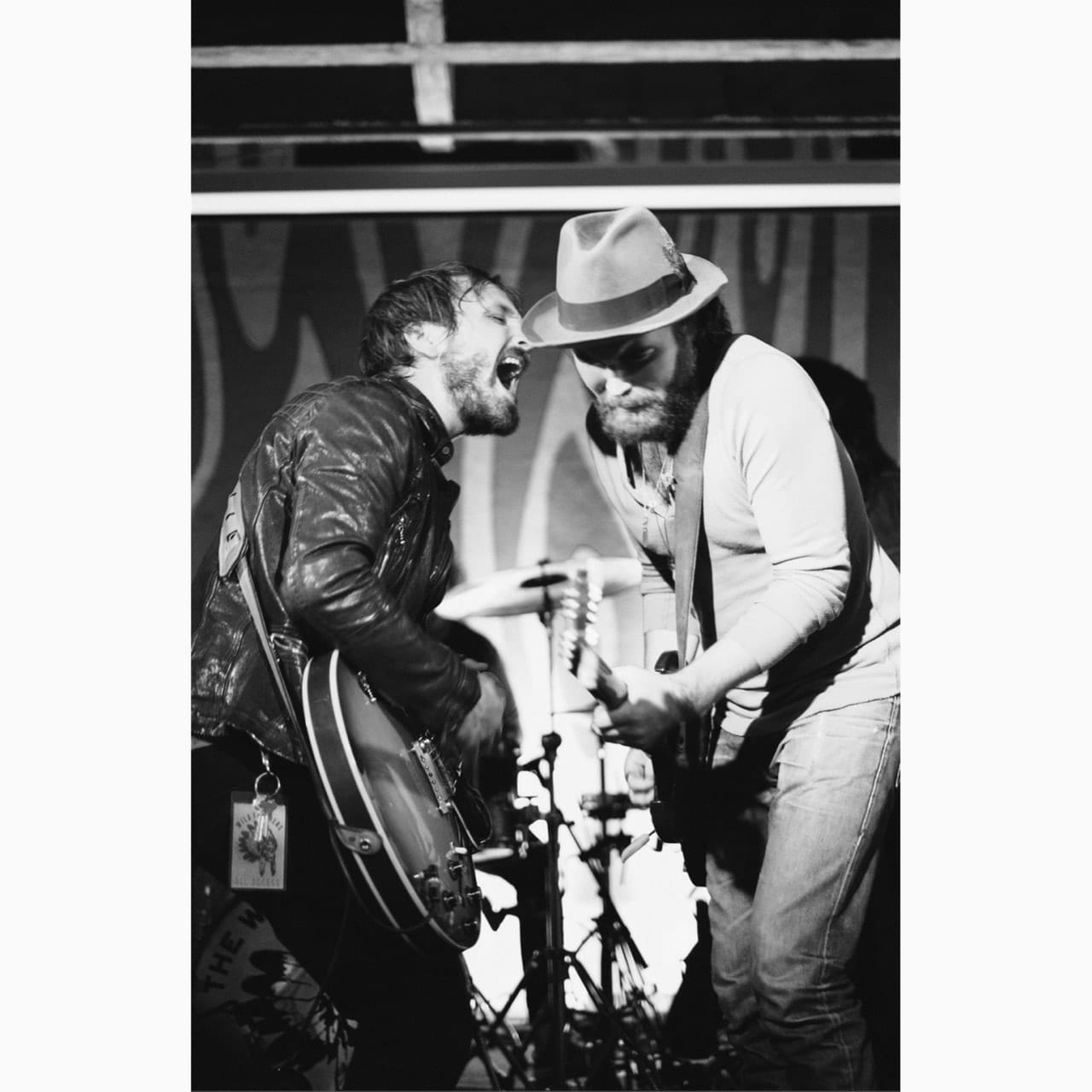 Black-and-white live photograph of two unidentified Wild Feathers guitarists leaning toward each other onstage.