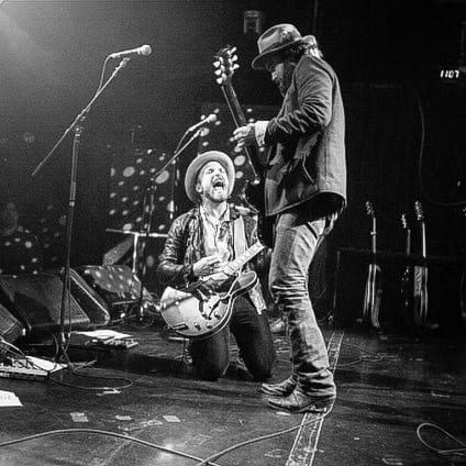 Black-and-white photograph of two unidentified Wild Feathers guitarists performing, one kneeling and one standing.