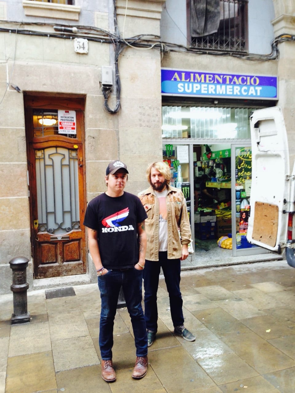 Two unidentified Wild Feathers members standing on a wet street outside a shop marked Alimentació Supermercat.