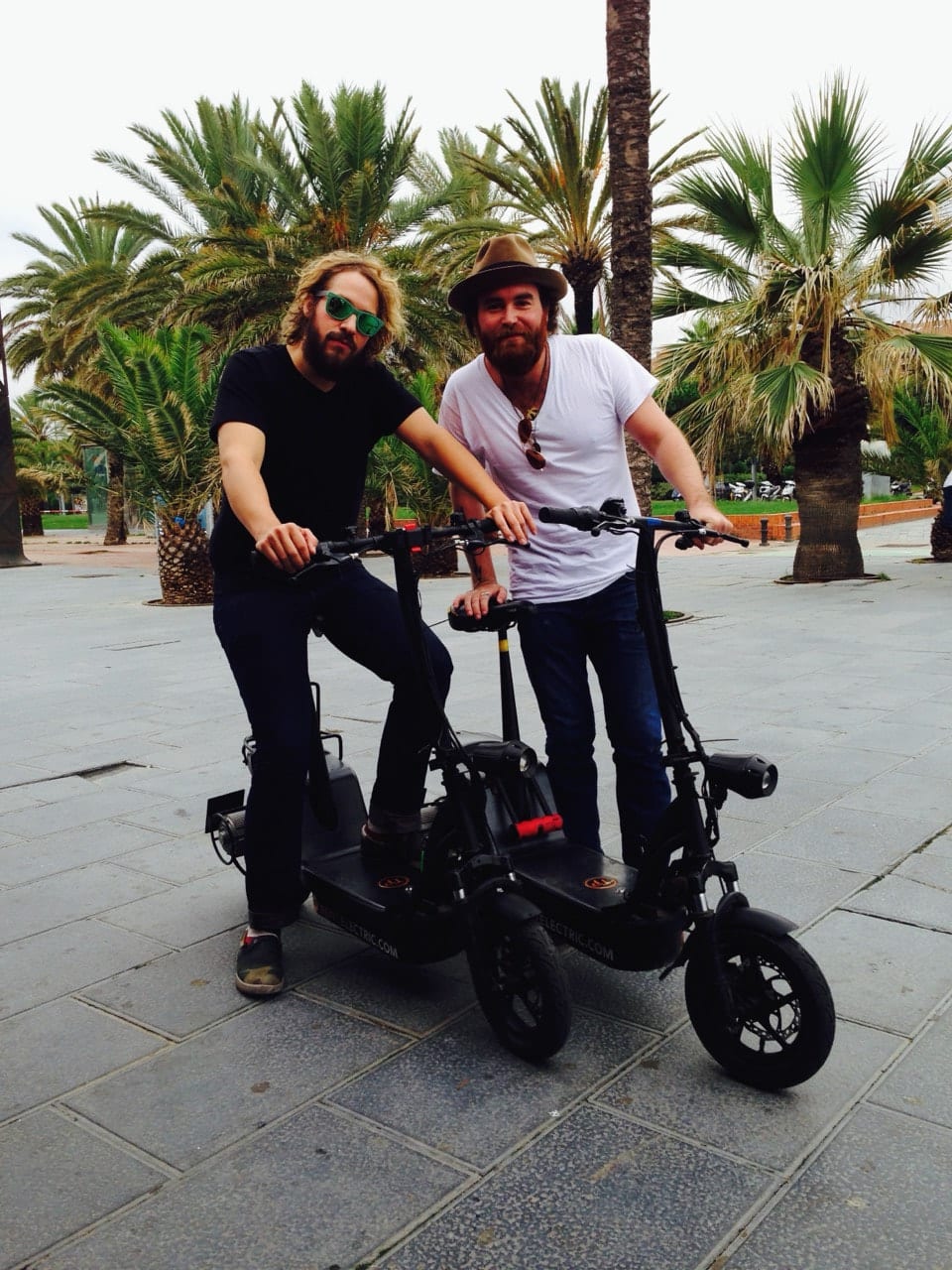 Two unidentified Wild Feathers members posing with compact electric scooters among palm trees during a probable Barcelona visit.