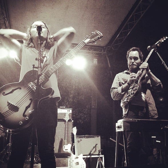 Two unidentified Wild Feathers members onstage with a hollow-body bass, guitar, microphones, and amplifiers.