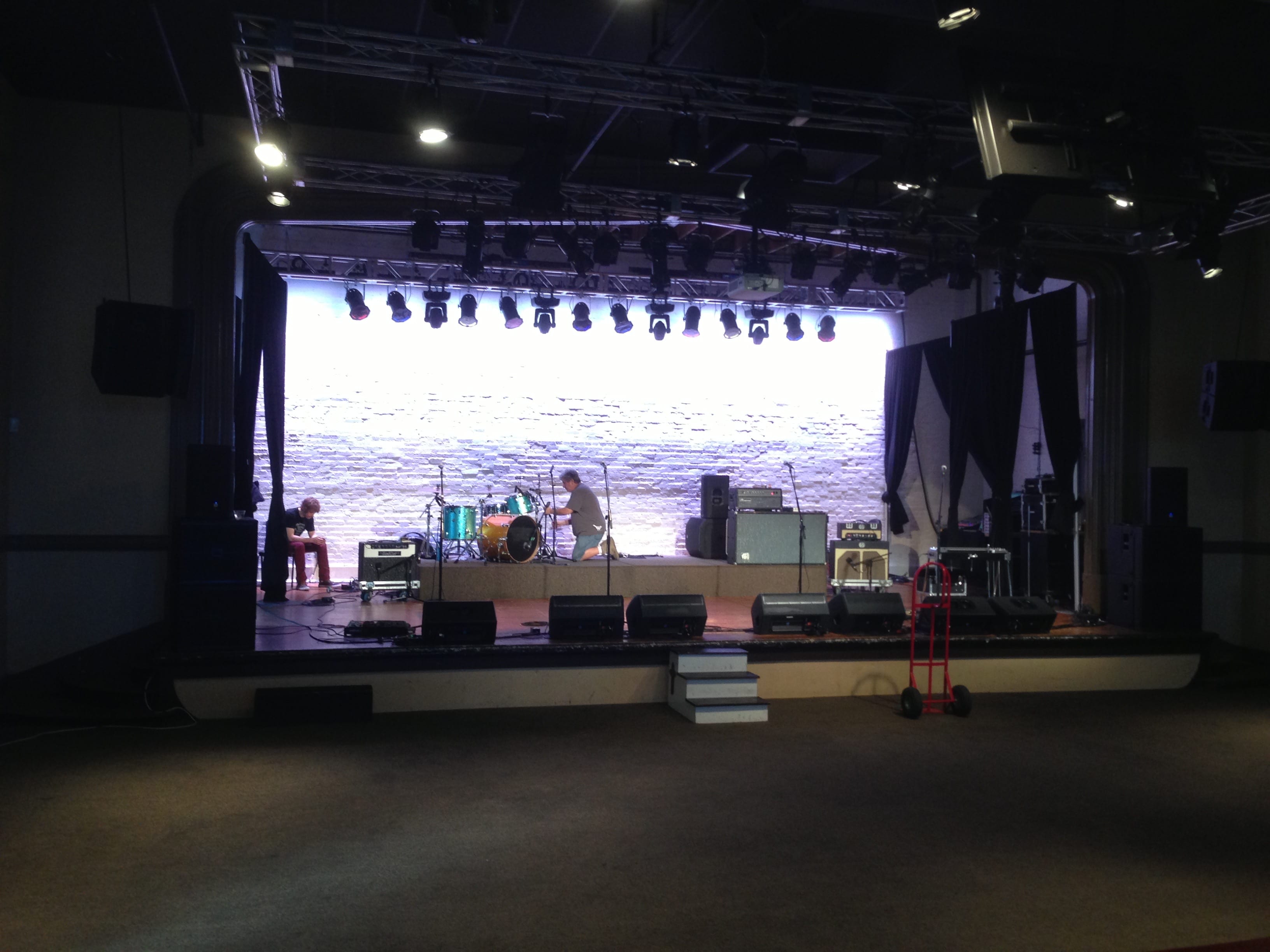 An empty concert room during load-in, with two people working on a stage holding drums, amplifiers, microphone stands, monitors, and a red hand truck.