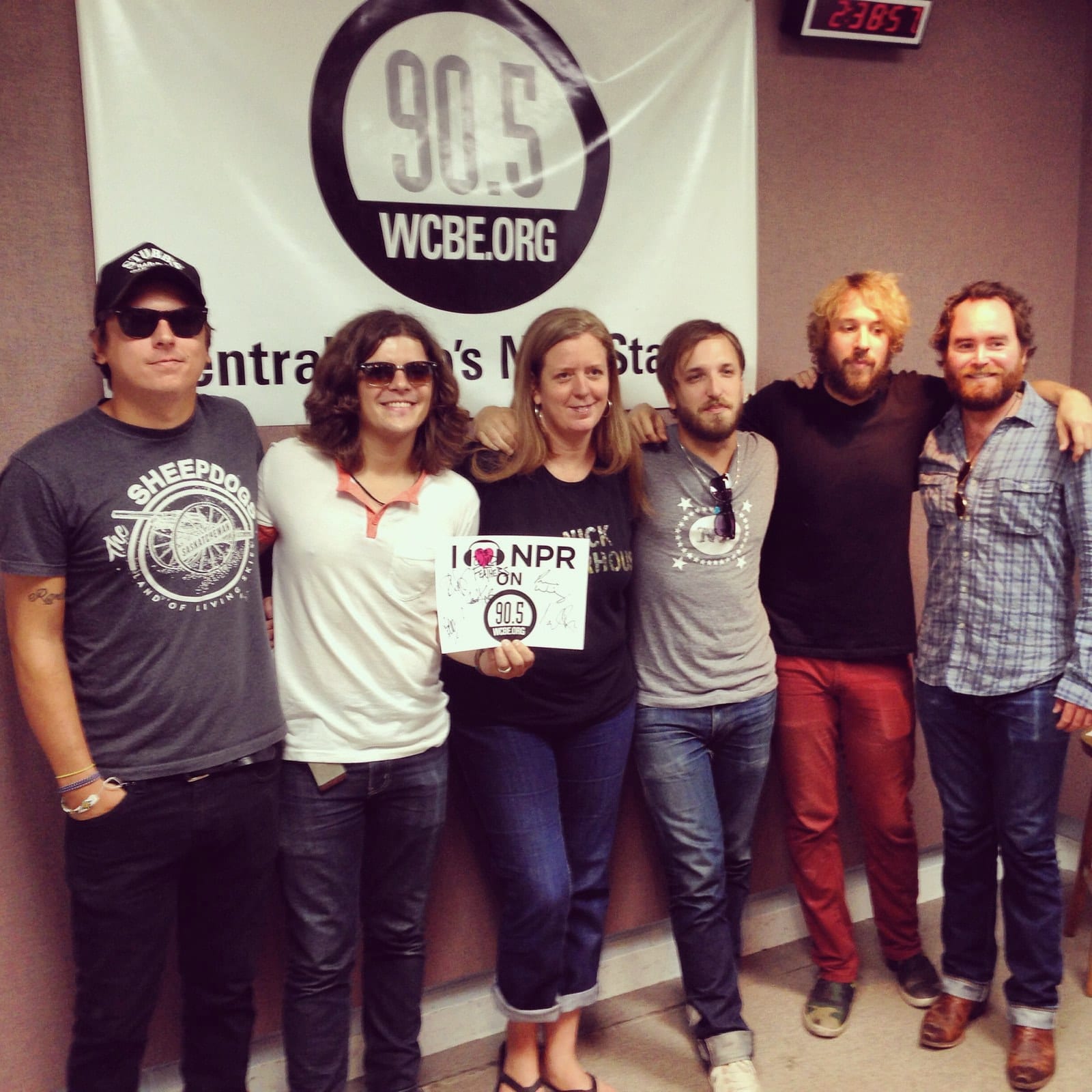 Members of The Wild Feathers and radio staff posing in front of a 90.5 WCBE sign in Columbus.