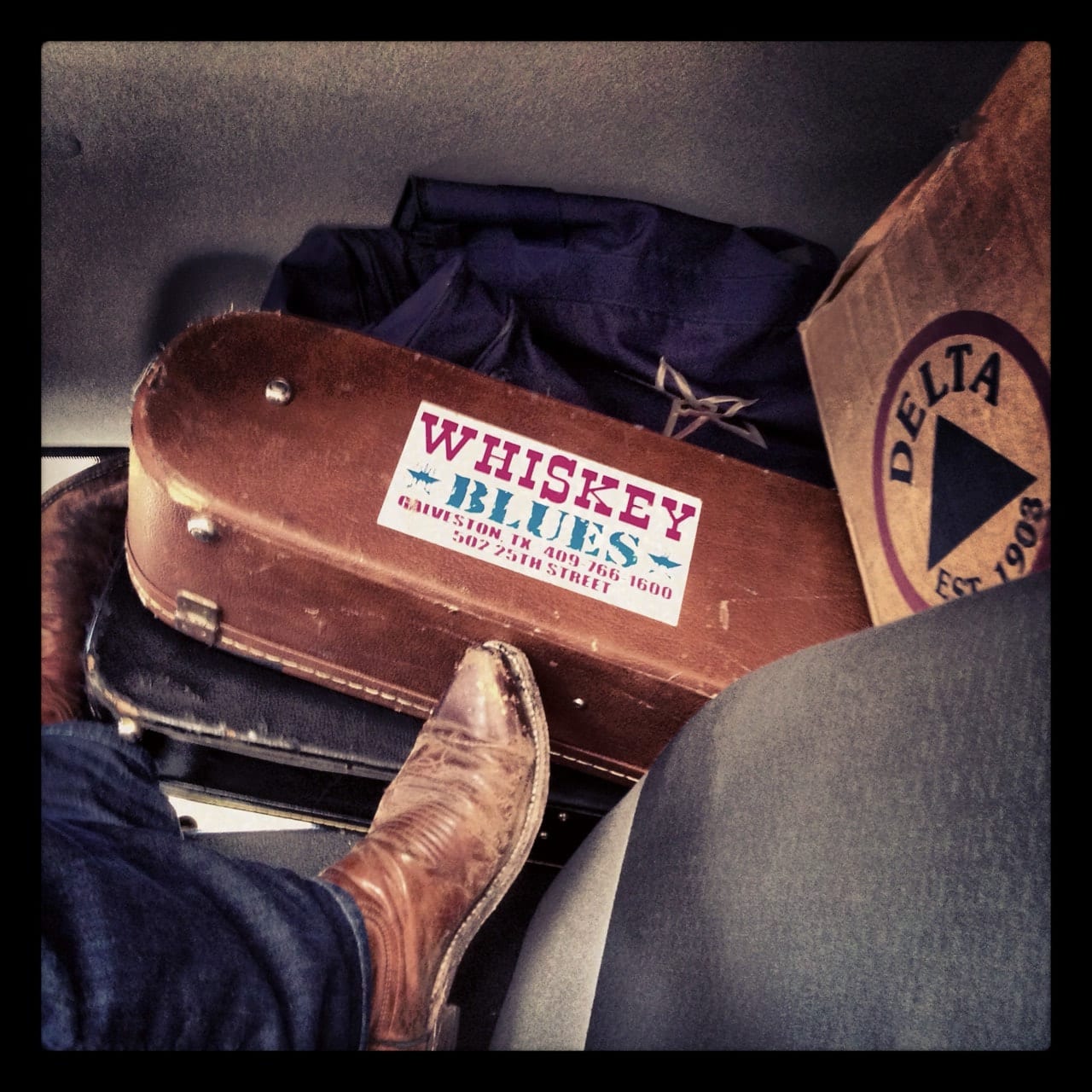 A boot resting against stacked instrument cases and luggage, including a case with a Whiskey Blues Galveston sticker and a Delta-branded box.
