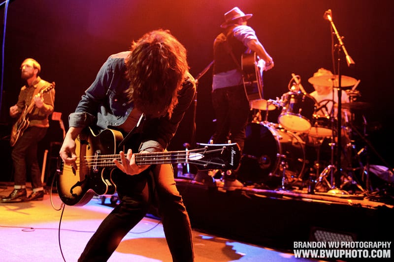 The Wild Feathers performing under red stage light at the 9:30 Club, with a bassist leaning into the foreground and three bandmates behind him.